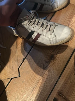 Vita Moncler sneakers med ränder - Snygga vita sneakers från Moncler med klassiska gröna och röda ränder på sidan samt Moncler-logga på plösen. Skorna har låga skaft, vita snören och sula i gummi med Moncler-mönster undertill. Tillverkade i läder för en clean och stilren look.