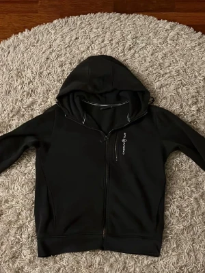 Svart Sail Racing hoodie - Säljer en svart hoodie från Sail Racing med dragkedja framtill och en liten logga på bröstet. Hoodien har huva och diskreta vita detaljer vid halsen. Perfekt för dig som gillar stilrena och sportiga plagg.