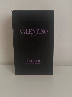 Valentino Uomo Born In Roma parfym - Valentino Uomo Born In Roma Purple Melancholia är en exklusiv parfym i en elegant svart kartong med lila text. Doften är modern och unik, perfekt för dig som vill sticka ut. Flaskan är troligtvis av glas och designen känns lyxig och trendig.