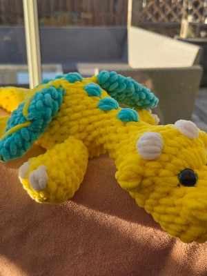 Gul virkad drakmjukis - Supergullig handvirkad drake i mjukt gult garn med turkosa detaljer på ryggen och vita horn. Draken har stora säkerhets ögon och är perfekt som dekoration eller mysig kompis. Unik och färgglad design som verkligen sticker ut.