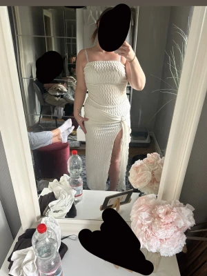 Vit långklänning med slits och knyt - Trendig vit långklänning med snygg slits framtill. Klänningen har ett strukturerat tyg och ett dekorativt knyt i midjan med metallspänne. Perfekt för sommarens alla tillfällen.