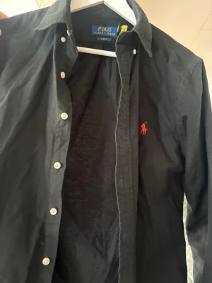 Svart skjorta från Polo Ralph Lauren - Klassisk svart skjorta från Polo Ralph Lauren med röd broderad logga på bröstet. Slim fit-modell med vita knappar och lång ärm. Tillverkad i mjuk bomull som känns skön mot huden. Perfekt för dig som gillar stilrena och tidlösa plagg.