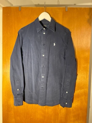 Mörkblå skjorta från Polo Ralph Lauren - Klassisk mörkblå skjorta från Polo Ralph Lauren med vit broderad logga på bröstet. Skjortan har långa ärmar, knappar framtill och på manschetterna samt en stilren krage. Perfekt för en clean och tidlös look.