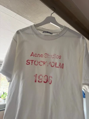Vit Acne Studios t-shirt Stockholm - Vit t-shirt från Acne Studios med röd text på bröstet där det står 'Acne Studios STOCKHOLM 1996'. Klassisk rund halsringning och korta ärmar. Tillverkad i mjuk bomull, perfekt för en clean och stilren look.
