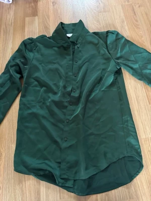 Mörkgrön skjorta i satin från Lindex - Snygg mörkgrön skjorta från Vila i glansigt satinmaterial. Skjortan har klassisk krage, knäppning framtill och långa ärmar. Perfekt för dig som vill ha en stilren och trendig look.