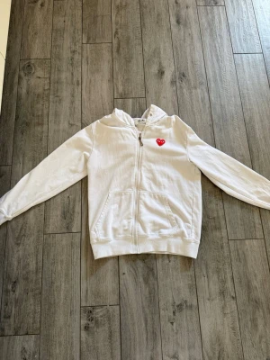 Vit hoodie från Comme des Garçons - Säljer en vit hoodie med dragkedja från Comme des Garçons Play. Två fickor framtill och ribbade muddar. Tillverkad i mjuk bomull och har en klassisk huva. Perfekt för en clean och stilren look. Passar perfekt som S också!