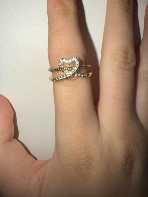 Guld ring med hjärta och stenar - Snygg guldfärgad ring med ett hjärta i mitten, dekorerad med gnistrande små stenar. Ringen har dubbla band och en öppen baksida som gör den justerbar. Perfekt accessoar för dig som gillar detaljer och bling.