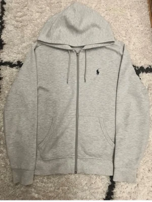 Grå hoodie från Polo Ralph Lauren - Snygg ljusgrå hoodie med dragkedja från Polo Ralph Lauren. Klassisk design med huva och dragsko, två fickor framtill och den ikoniska broderade loggan på bröstet. Perfekt för en chill och stilren look.