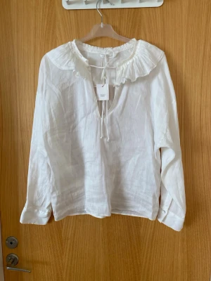 Vit linnéblus med volangkrage Arket - Vit blus från Arket i 100% linne med lång ärm, volangkrage och knytband vid halsen. Blusen har en avslappnad passform och lätt struktur i tyget, perfekt för en trendig och luftig stil.