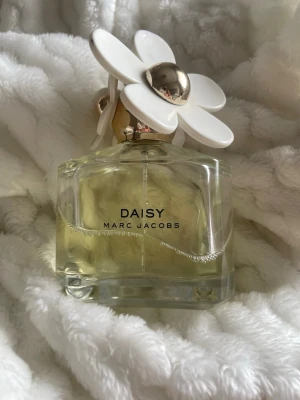 Daisy parfym från Marc Jacobs - Daisy från Marc Jacobs är en ikonisk parfym i en genomskinlig glasflaska med gulaktig vätska. Locket är dekorerat med stora vita blommor och guldfärgade detaljer, vilket ger en lekfull och lyxig känsla. Perfekt för dig som gillar snygg design och fräscha dofter. Något använd, se bild. Tyvärr passar inte doften mig. Den ena guld knoppen är delvis borta efter en damning. 