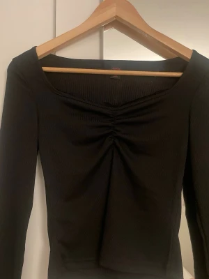 Svart ribbad topp med rynk framtill - Trendig svart långärmad topp från Monki med ribbad struktur och snygg rynkdetalj framtill. Toppen har en bred, något fyrkantig ringning och sitter figurnära. Perfekt att styla med både jeans och kjol.