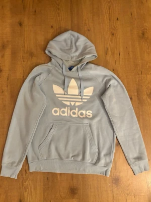 Ljusblå hoodie från Adidas - Snygg ljusblå hoodie från Adidas med stor vit Trefoil-logga och text på bröstet. Klassisk känguruficka framtill och justerbar huva med dragsko. Tillverkad i mjukt material som känns skönt mot huden. Perfekt för en avslappnad streetwear-look.