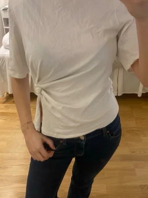 Vit topp med knyt i sidan - Säljer en vit topp med korta ärmar och snygg knytdetalj i sidan som ger en cool twist. Toppen har rund halsringning och är gjord i mjukt bomullsmaterial. Perfekt till jeans eller kjol för en enkel men trendig look.