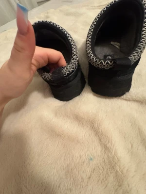 Svarta UGG tofflor med mönstrad kant - Säljer ett par svarta UGG tofflor med gråvit mönstrad kant runt öppningen. Tofflorna har en mjuk och fluffig insida och är perfekta för att hålla fötterna varma. Klassisk slip-on modell med robust sula och UGG-logga på innersulan. RIKTIGA UGGS!!!