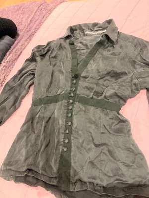 Grågrön blus med knappar - Stilren grågrön blus med långa ärmar och dekorativa knappar framtill. Blusen har markerad midja med gröna banddetaljer och klassisk krage. Perfekt till jeans eller kjol för en trendig look.