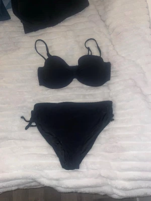 Svart bikini från Blue Lovers - Snygg svart bikini från Blue Lovers med vadderad bygel-bh och justerbara axelband. Bikinitrosan har hög midja och knyt i sidorna för en trendig look. Perfekt för strand eller poolhäng i sommar!