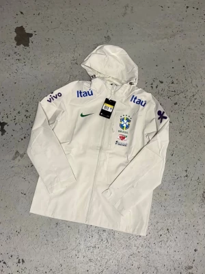 Vit Brasilien vindjacka med tryck - Snygg vit vindjacka från Nike med Brasilien-logga och flera sponsortryck i blått, lila och grönt. Jackan har huva, dragkedja framtill och tryck på bröst och ärmar. Perfekt för dig som gillar sportig stil och vill sticka ut.