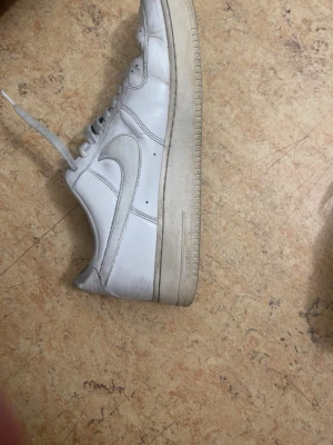Vita Nike Air Force 1 sneakers - Klassiska vita Nike Air Force 1 sneakers i lågt utförande. Skorna har en ikonisk swoosh-logga på sidan, perforerad tåbox för ventilation och en kraftig sula. Tillverkade i läder med textilfoder och snörning framtill. Perfekta för en clean och tidlös streetstil.