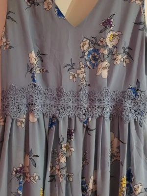 Blommig blå långklänning med spets - Långklänning i ljusblått med blommigt mönster i vitt, blått och gult. Klänningen har v-ringning, spetsdetalj i midjan och är ärmlös. Perfekt för sommarens alla tillfällen.