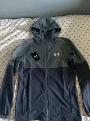 Grå vindjacka från Under Armour - Snygg tvåfärgad vindjacka från Under Armour i mörkgrått och ljusgrått. Jackan har huva, dragkedja framtill och två sidofickor. Perfekt för blåsiga dagar och sportiga outfits. Under Armour-logga på bröstet och detaljer på ärmen. Använd typ 2 gånger så de är väldigt bra skick. Bara skriva för fler bilder eller frågor 