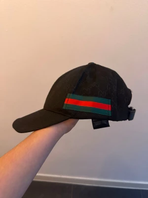 Svart keps från Gucc i med GG-loga  - Svart keps från Gucc i med klassiskt GG-monogram över hela tyget och en grön-röd rand på sidan. Justerbart spänne bak och böjd skärm. Tillverkad i ett slitstarkt tyg som känns lyxigt och stilrent.