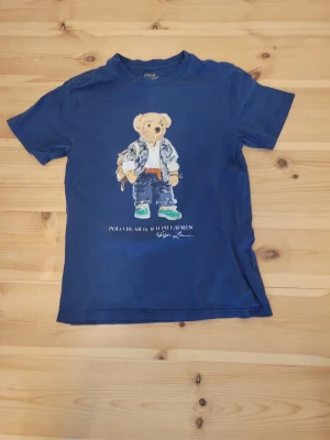 Blå Polo Ralph Lauren t-shirt med björn - Säljer en blå t-shirt från Polo Ralph Lauren med ikoniskt björntryck på bröstet. T-shirten har rund hals och korta ärmar. Tillverkad i mjuk bomull som är skön att bära. Perfekt för dig som gillar klassiska streetwear-vibbar.