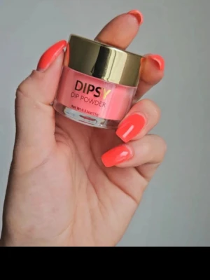 DIPSY Dip Powder i korall - Säljer ett DIPSY Dip Powder i en snygg korall nyans. Kommer i en rund burk med guldfärgat lock och innehåller 15g pulver. Perfekt för att skapa trendiga och hållbara naglar med en fräsch färg. Pulvret är lätt att applicera och ger ett jämnt resultat.