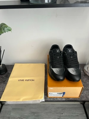 Svarta Louis Vuitton sneakers - Snygga svarta sneakers från Louis Vuitton med klassisk LV-logga på plösen och framtill. Skorna är låga och har en stilren design i läder med svarta skosnören. Perfekta för dig som vill ha en exklusiv och trendig look.