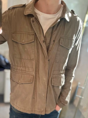 Beige fieldjacket - Stilren fieldjacket med fyra stora fickor framtill och dragkedja. Jackan har klassisk krage, axelklaffar och är tillverkad i mjuk bomull. Perfekt för lager på lager och enkel att matcha med olika outfits.