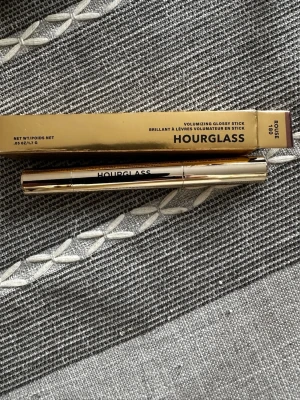 Hourglass Volumizing Glossy Stick Rouge - Snyggt läppglans i pennformat från Hourglass i nyansen Rouge. Kommer i en lyxig guldfärgad hylsa och ger en glansig finish med volym till läpparna. Perfekt för dig som vill ha både färg och glans i ett smidigt format.