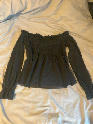 Svart offshoulder blus med långa ärmar - Trendig svart offshoulder blus med smockad överdel och långa, luftiga ärmar. Blusen har en avslappnad passform och volangkant upptill. Perfekt att styla med jeans eller kjol för en cool look.