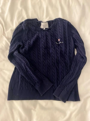 Mörkblå kabelstickad tröja Bondelid - Säljer en mörkblå kabelstickad tröja från Bondelid med rund halsringning och diskret broderad logga på bröstet. Tröjan har långa ärmar och ribbade muddar vid ärmslut och nederkant. Perfekt för dig som gillar klassisk och stilren look.