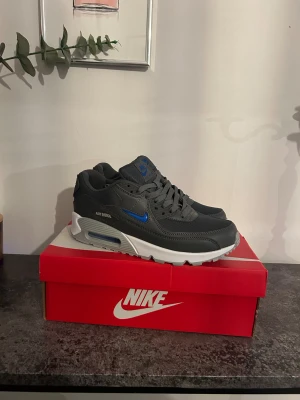 Nike Air Max 90 mörkgrå/blå sneakers - Nike Air Max 90 sneakers i mörkgrått med blå detaljer och vit/grå sula. Klassisk siluett med snörning och synlig Air Max-dämpning. Ovandel i syntet och mesh för både stil och komfort. Perfekta för dig som gillar streetwear och sportig vibe.