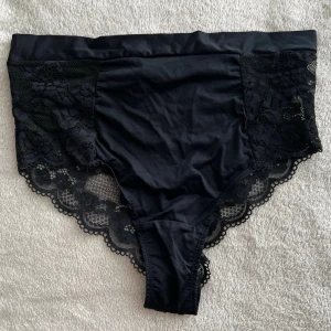 Svarta spetsdetaljerade trosor - Ny pris 120kr. Säljer ett par svarta trosor med bred spetskant och blommönstrad spets på sidorna och baktill. Trosorna har hög midja och är tillverkade i mjukt, stretchigt material som ger en bekväm passform. Perfekta för dig som gillar en elegant och feminin stil.