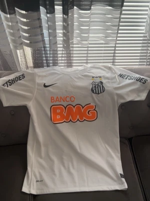 Vit Santos Neymar Jr matchtröja - Säljer en vit Santos matchtröja från Nike med Neymar Jr och nummer 11 på ryggen. Tröjan har tryckta sponsorer som BMG och Netshoes, samt klubbmärke på bröstet. Tillverkad i lätt och ventilerande material, perfekt för fotboll eller samling.