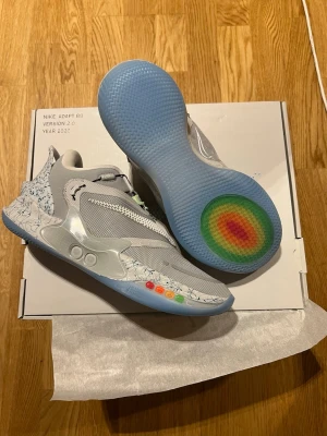 Nike Adapt BB 2.0 Grey Sneakers - Sjukt futuristiska Nike Adapt BB 2.0 sneakers i grått med självknäppande teknologi, mesh och syntetmaterial, genomskinlig sula med regnbågsdetalj och färgglada knappar på sidan. Swoosh-logga på ovansidan och coolt mönster på hälen. Aldrig använda och allt finns med i lådan. Finns ej att köpa så dessa är väldigt unika.