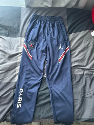 PSG x Jordan blå mjukisbyxor - Snygga blå mjukisbyxor från PSG x Jordan med vita och röda detaljer längs benen. Byxorna har elastisk midja, PSG-logga och Jumpman-logga framtill samt texten PARIS på ena benet. Perfekta för en sportig och avslappnad stil.