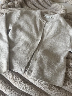 Ljusbeige kofta från Kappahl - Mysig ljusbeige kofta från Kappahl i mjukt stickat material. Rund halsringning, knäppning framtill med små knappar och ribbade muddar vid ärmslut och nederkant. Perfekt för lager på lager och enkel att matcha med olika outfits.