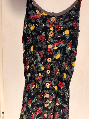 Svart Deadpool-klänning med knappar - Cool svart klänning med Deadpool-tryck, tropiska blommor och blad i rött, gult och grönt. Klänningen har träknappar framtill och smala axelband. Perfekt för dig som gillar Marvel och vill sticka ut med färgglatt mönster.