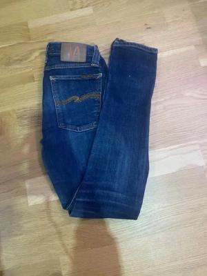 Mörkblå jeans från Nudie Jeans - Snygga mörkblå jeans från Nudie Jeans med klassisk femficksmodell och gul kontrastsöm på bakfickan. Jeansen har en rak passform och är tillverkade i slitstark denim. Perfekta för dig som gillar stilrena och tidlösa jeans.