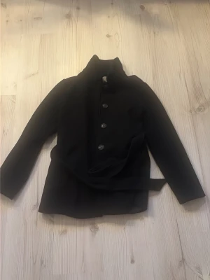 Svart kappa från Vila med skärp - Stilren svart kappa från Vila i storlek 36. Kappan har knäppning framtill med fem knappar skriv om du vill se hur den ser ut på!