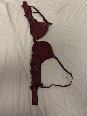 Vinröd spets-bh med vaddering - Snygg vinröd bh med vadderade kupor och detaljer i spets. Bh:n har justerbara axelband och hakknäppning bak. Dekorativ liten rosett mellan kuporna. Perfekt för dig som gillar romantiska underkläder med fina detaljer.