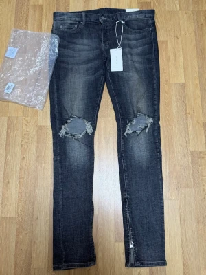 Säljer ett par MNML Ripped Stretch Jeans Grey – Size W32 - Jeansen är helt nya och endast testade. Inga skador eller slitage.  Trendiga jeans i grå tvätt med riktiga ripped/hål vid knäna, vilket ger en tydlig streetwear-look. Dragkedjor vid anklarna för extra stil. Original tags medföljer.  Size: W32 🏷️ Rabatt vid köp av flera plagg!
