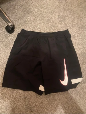 Svarta Nike shorts med vit logga - Säljer ett par svarta Nike shorts med stor vit Swoosh-logga på vänster ben och vita detaljer längst ner på båda benen. Shortsen har elastisk midja och är perfekta för sport eller chill. Klassisk och sportig look.