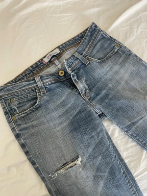 Blå bootcut jeans - Säljer ett par blå bootcut jeans med öppningar på låret och knät. Jeansen har låg midja och klassisk femficksdesign. Innerbenslängd: 74cm och jag är 162cm, står inte vilken storlek det ör på byxorna men måtten är på bild 3-6. pris går att diskutera❣️