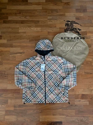 Burberry Windbreaker - 🌬️ Burberry Reversibel Windbreaker – Nyskick 🌬️ Stilren Burberry windbreaker i nyskick, reversibel med klassisk beige rutdesign på ena sidan och svart på andra. Justerbar huva med dragsko, hel dragkedja och elastiska ärmslut ger perfekt passform. Lätt och slitstarkt material gör jackan idealisk för alla väder. Två stilar i ett – elegant och funktionell. Nyskick med tagg kvar. 📦 Snabb leverans – kontakta vid frågor!