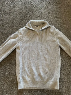 Ljusgrå stickad tröja från Zara - Snygg ljusgrå stickad tröja från Zara med halv dragkedja och hög krage. Tröjan har ribbstickad struktur och långa ärmar, perfekt för kyligare dagar. Enkel och stilren design som passar till det mesta. (Använt 2-3 gånger!)