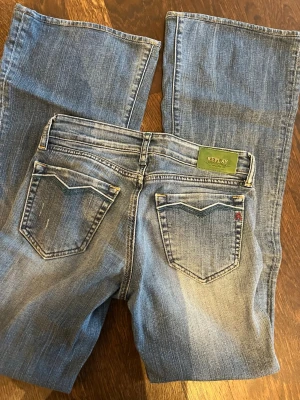 Blå bootcut jeans från Replay - Snygga blå bootcut jeans från Replay med klassisk femficksmodell och slitna detaljer. Jeansen har en ljus tvätt och dekorativa sömmar på bakfickorna. Tillverkade i bomull med en skön passform och detaljer som Replay-logga på bakfickan.