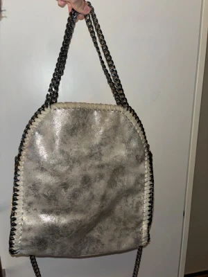 Silvrig axelväska med kedjeremmar - Cool axelväska i metallic-silver med kedjeremmar och dekorativa sömmar längs kanterna. Väskan har en mjuk, lite slouchy form och är tillverkad i konstläder. Perfekt för dig som gillar en edgy och trendig stil.