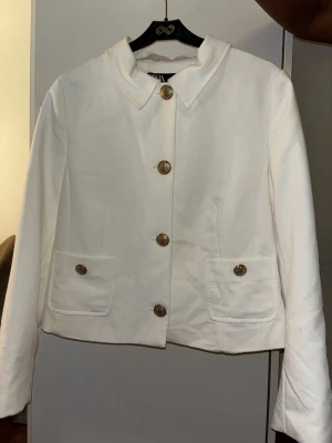 Vit kavaj med guldknappar - Stilren vit kavaj med fyra dekorativa guldknappar framtill och två fickor med lock. Kortare modell med mjuk krage och raka ärmar. Perfekt för att lyfta din outfit med en klassisk touch.